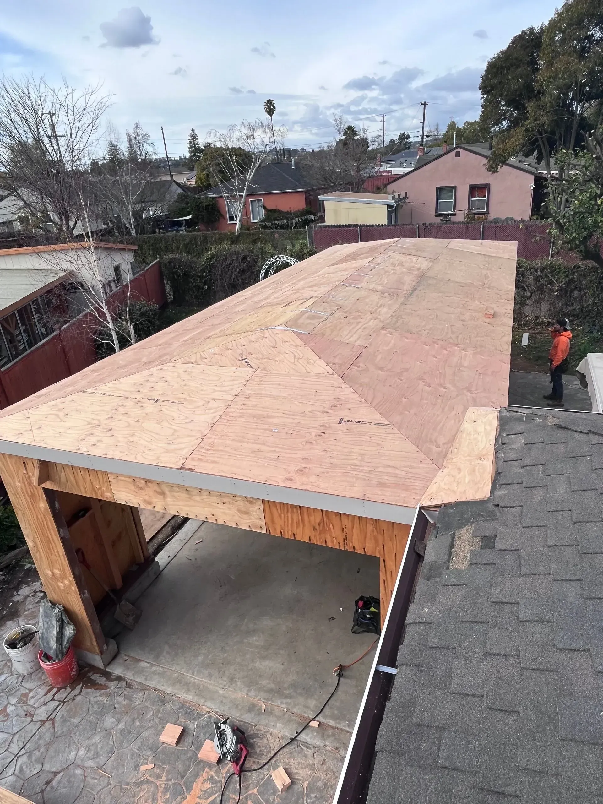 Garage roof construction in progress; plywood panels cover the framing; a worker stands nearby.