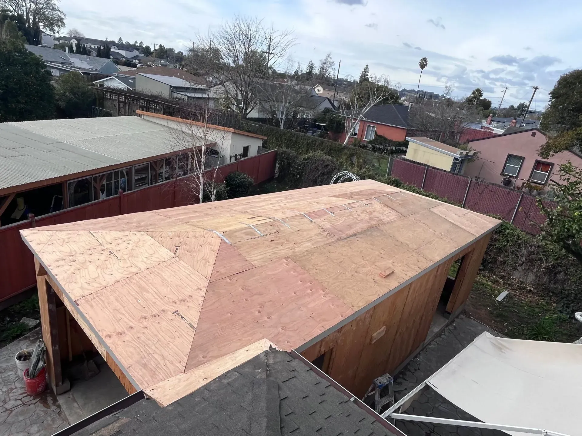 Newly constructed wooden shed with a partially completed plywood roof, surrounded by other houses and trees.