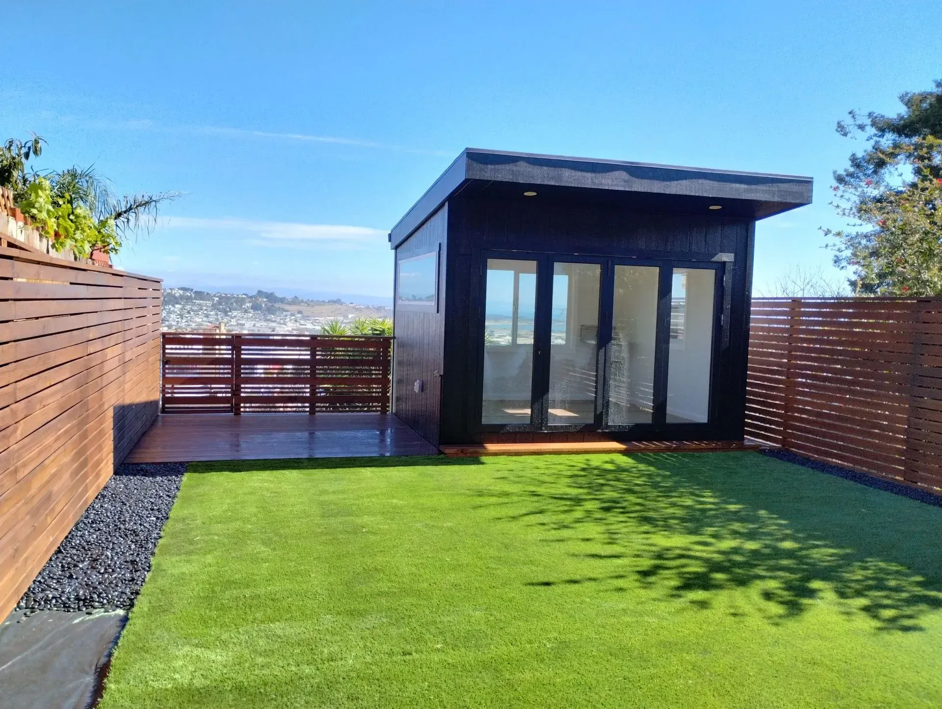 Black modern backyard office with glass doors, wooden deck, green lawn, and city view.