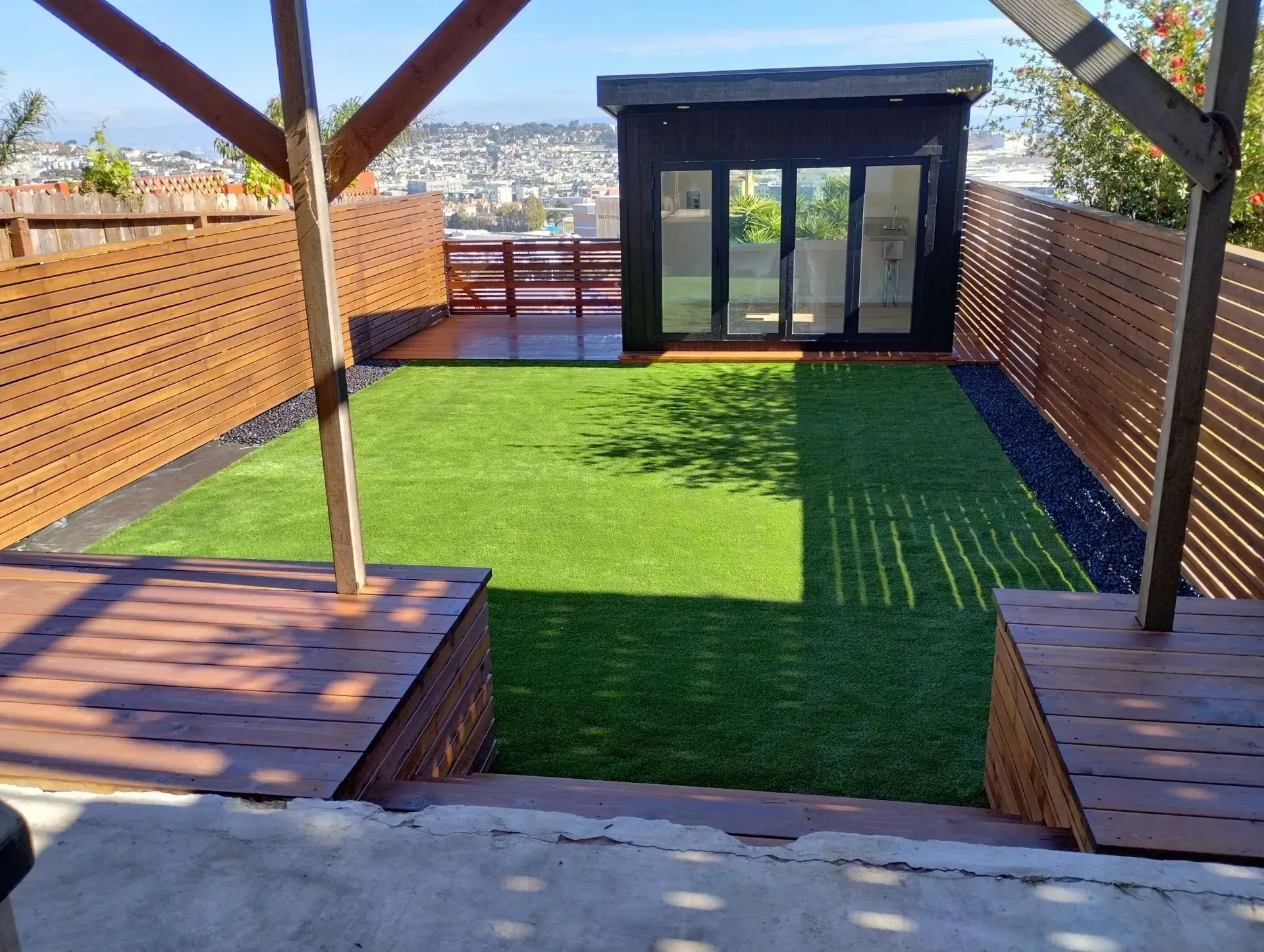 Backyard with green turf, wooden deck, and black shed with glass doors, overlooking a city.