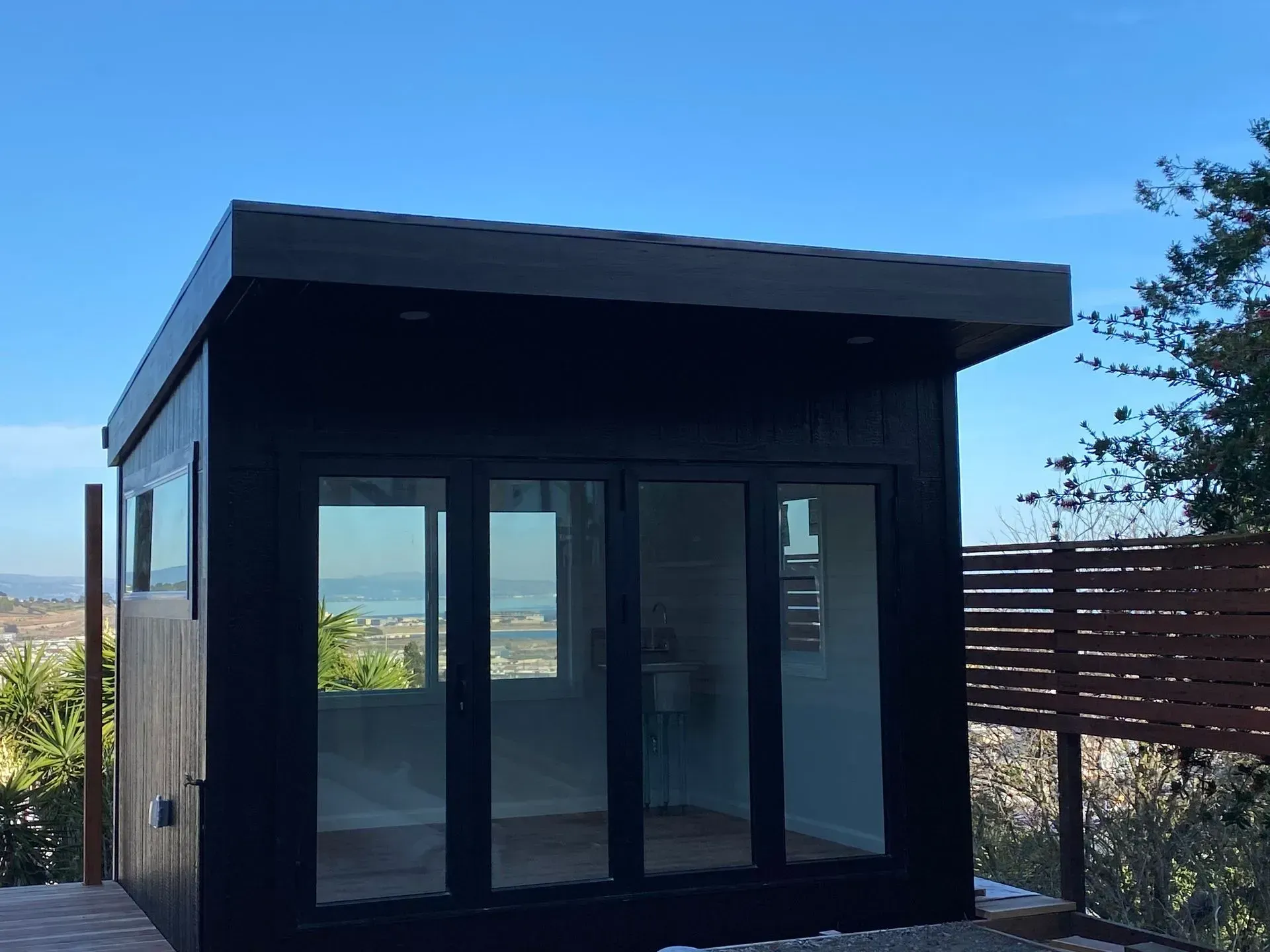 Black modern shed with glass doors and windows, brown roof, set against a cityscape backdrop.