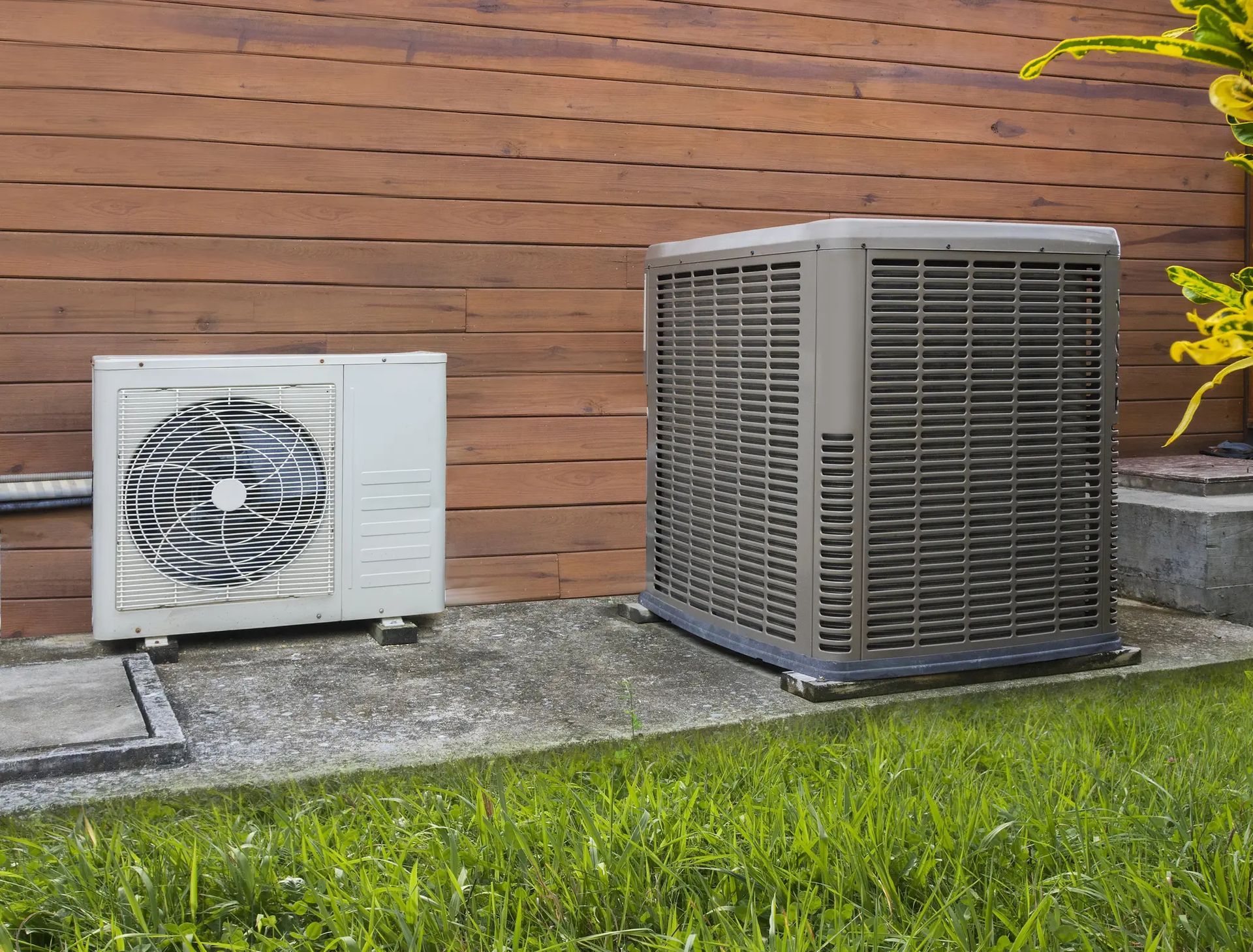 Two outdoor air conditioner units beside a wooden wall, one white and one gray, on a grassy lawn.