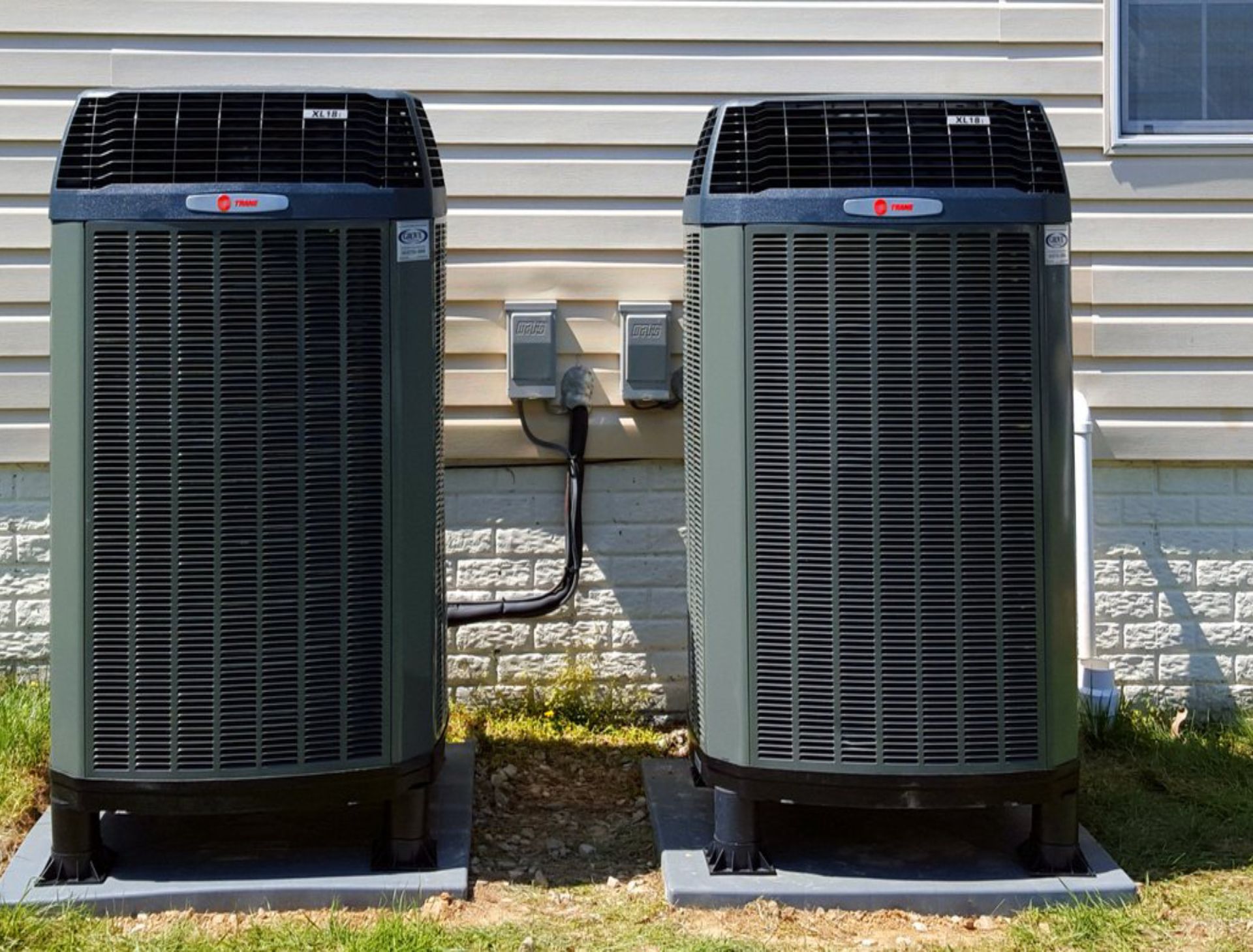 Two black outdoor air conditioning units beside a house wall on a grassy yard
