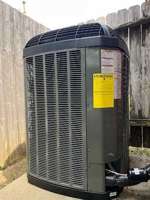 Outdoor gray HVAC unit beside a wooden fence and concrete wall, with a yellow warning label on the side