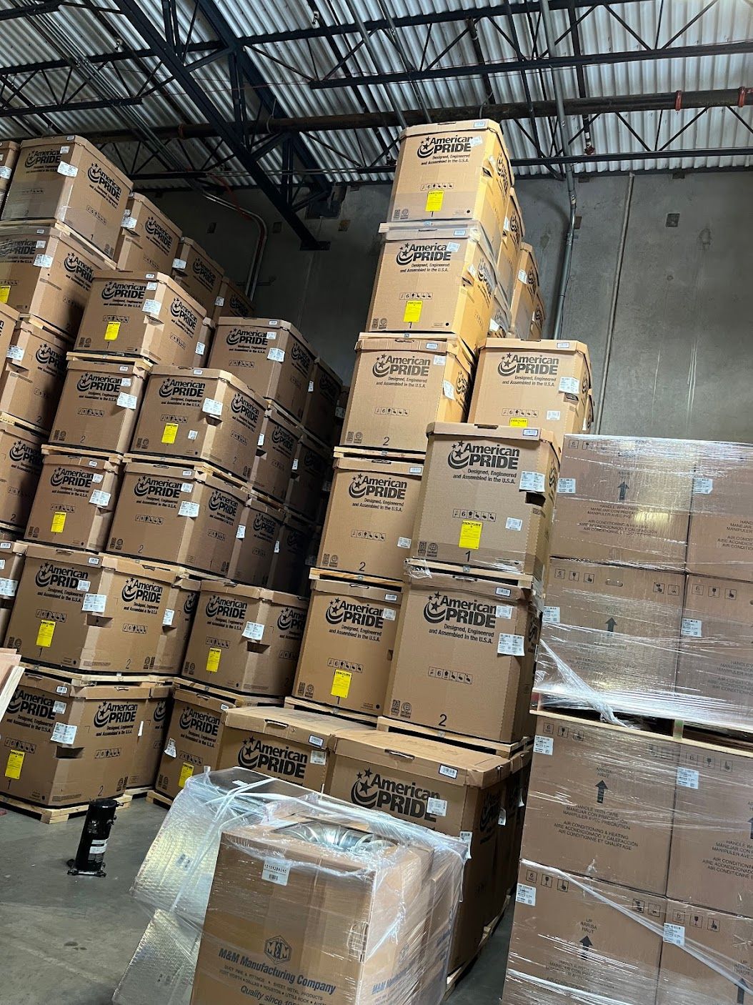 Stacks of taped cardboard boxes piled in a warehouse under a metal roof.