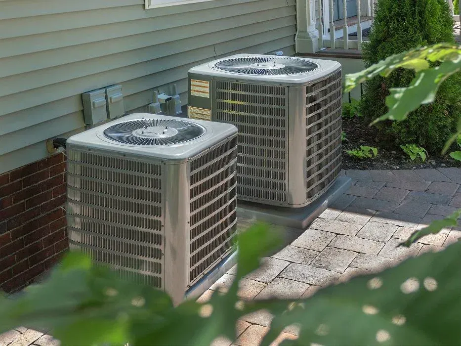 Two outdoor air conditioning units beside a house on a paved patio