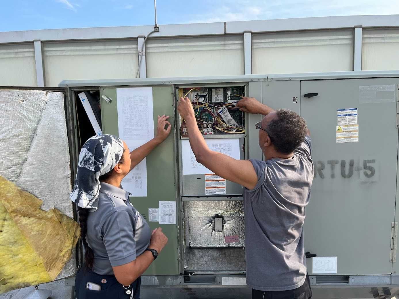 Two people working on exposed electrical components inside a gray utility box outdoors