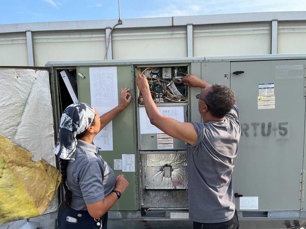 Two workers in gray shirts inspect electrical equipment inside a metal utility cabinet outdoors