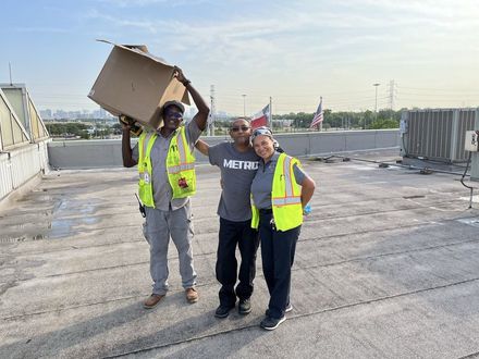 Three workers in safety vests stand on a rooftop, one holding a box overhead.