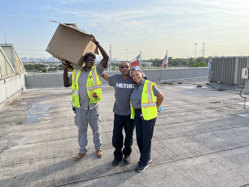Three workers in safety vests on a rooftop, one holding a box overhead and another waving.