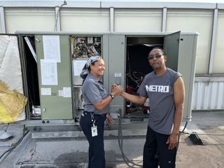Two people shaking hands beside an open electrical control panel outdoors.