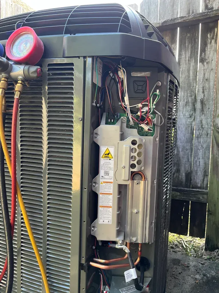 Open HVAC unit with exposed wiring, copper lines, and a red pressure gauge outdoors.