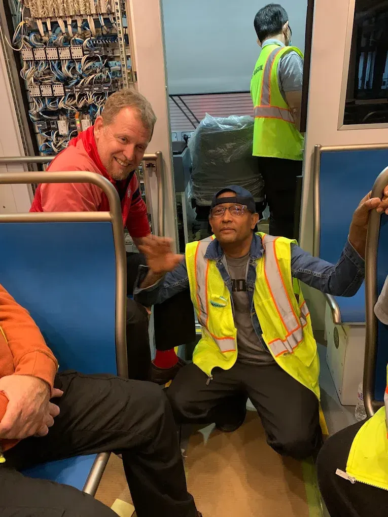 Passengers sitting on a train floor beside seats, wearing reflective safety vests, with one smiling and gesturing.