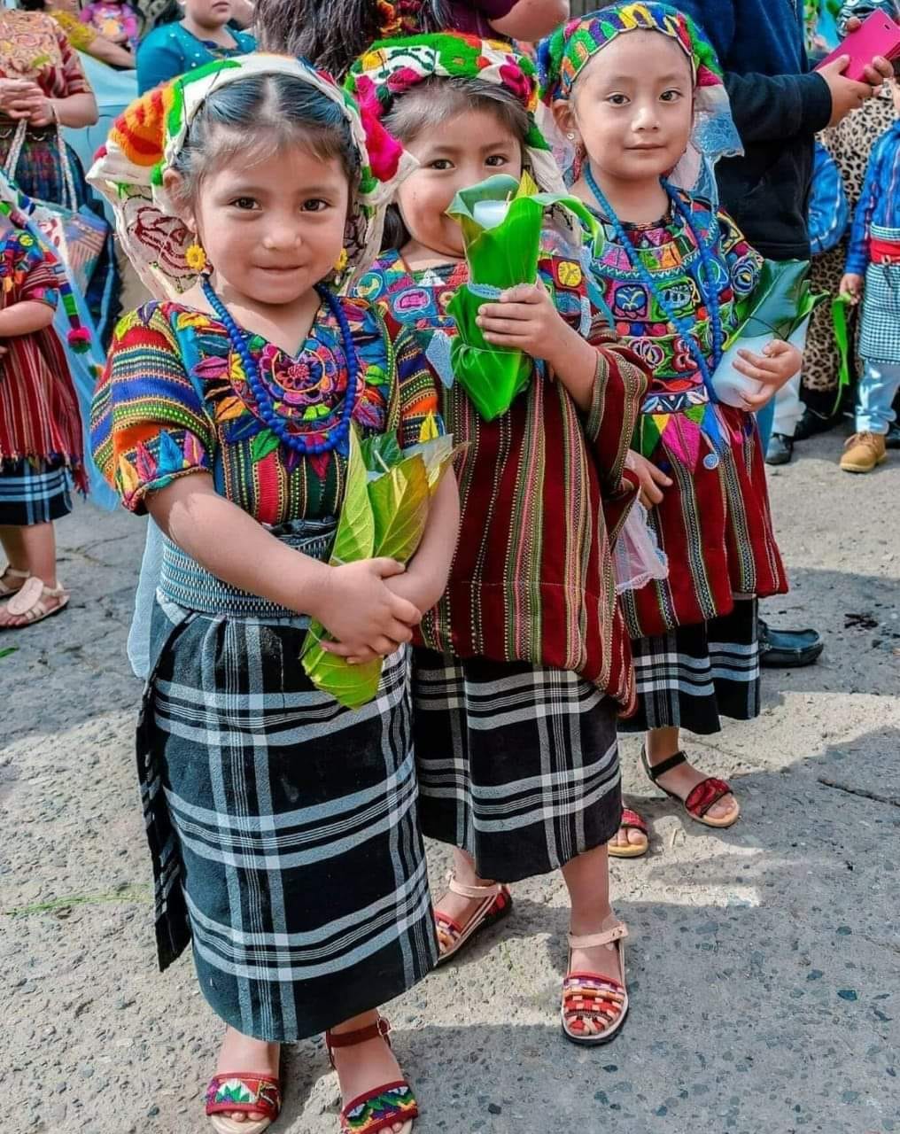 Drei Kinder in farbenfroher traditioneller guatemaltekischer Kleidung, die Geschenke halten und lächeln.
