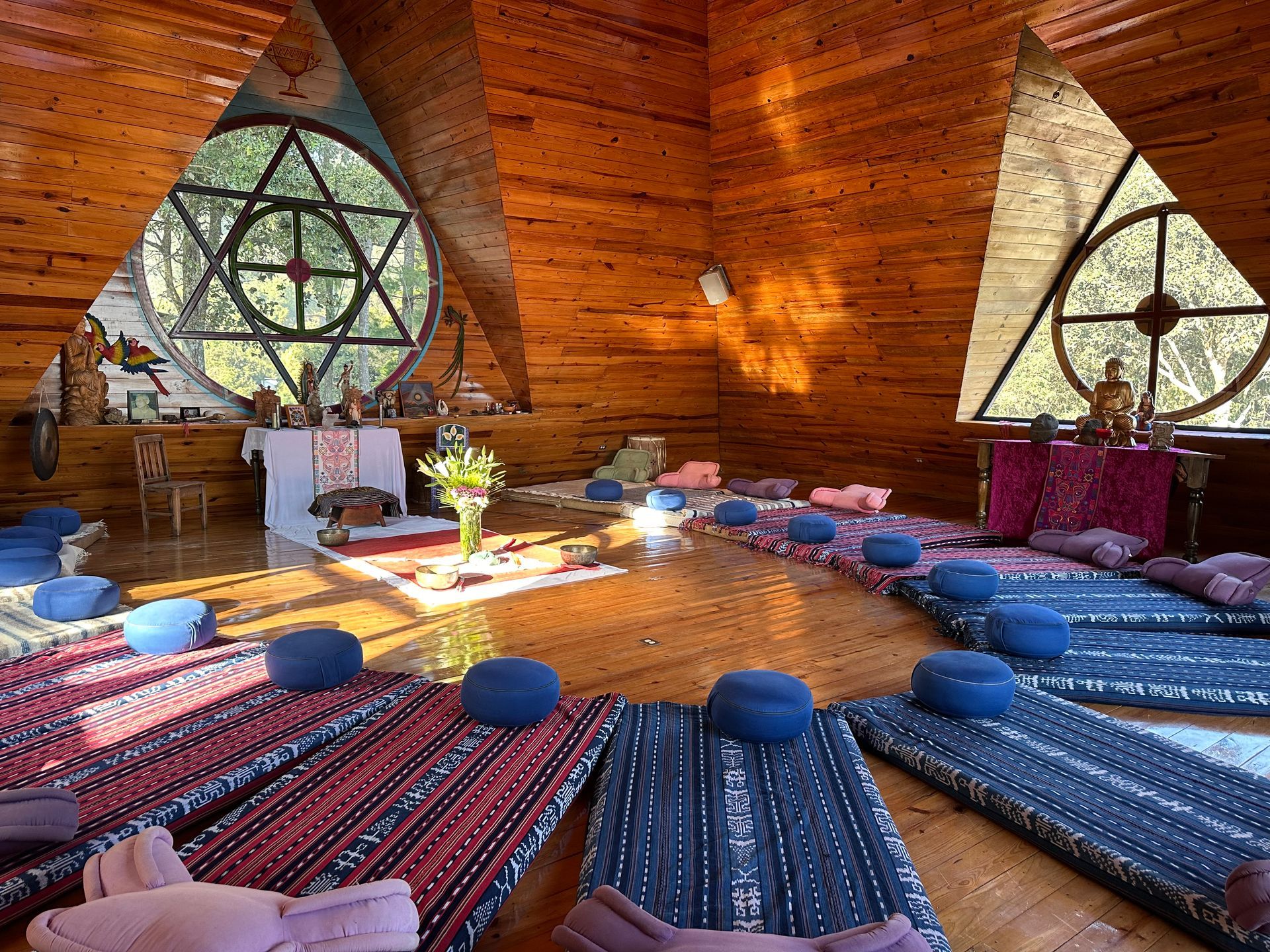 Meditationsraum mit im Kreis angeordneten Matten und Kissen, geometrischen Fenstern und holzgetäfelten Wänden.