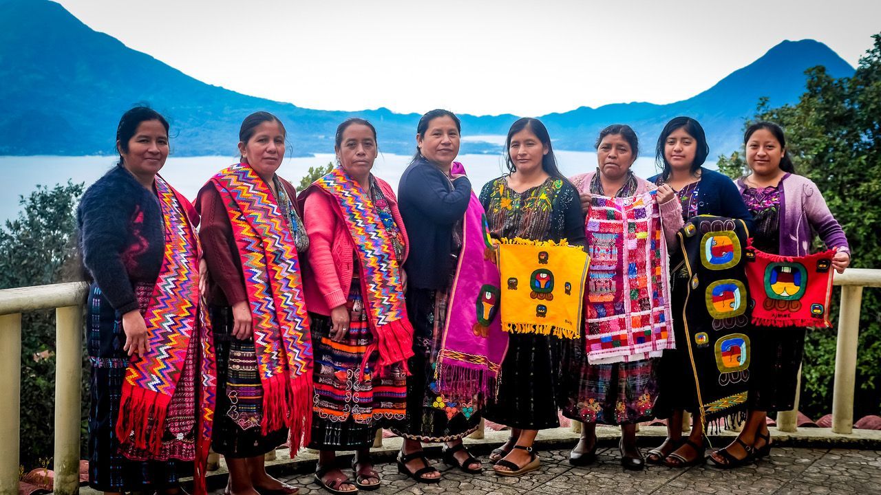 Eine Gruppe von Frauen in traditioneller Kleidung steht vor einem See und Bergen im Hintergrund.