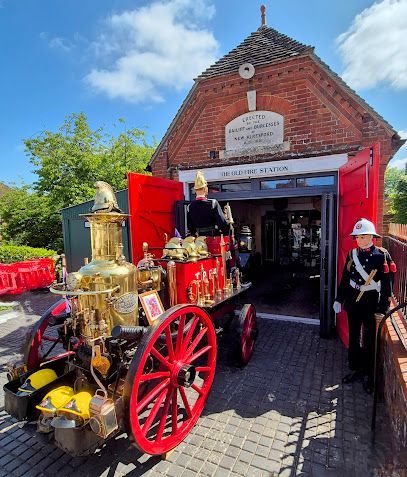 Red antique fire engine outside a brick museum building with open red doors and a firefighter statue.