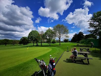 Golf course with green fairway, trees, blue sky, and people sitting at picnic tables near clubs