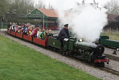 Miniature steam train pulling passengers through a park, with white steam billowing from the engine.