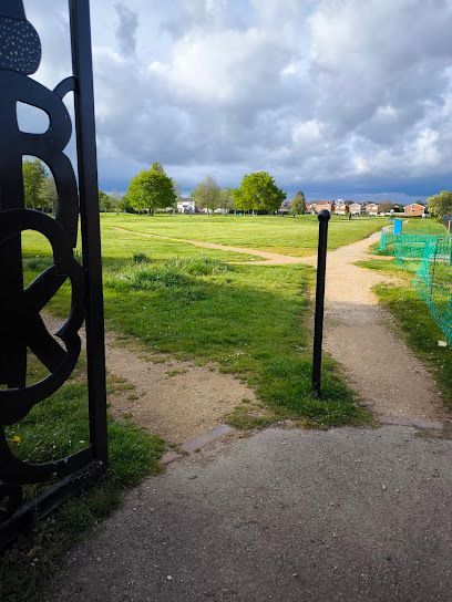 Grassy park path through an open gate under cloudy sky, with distant trees and blue fences