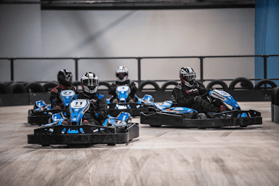 Four go-karts racing indoors, with blue and black karts leading on a wood-floored track.