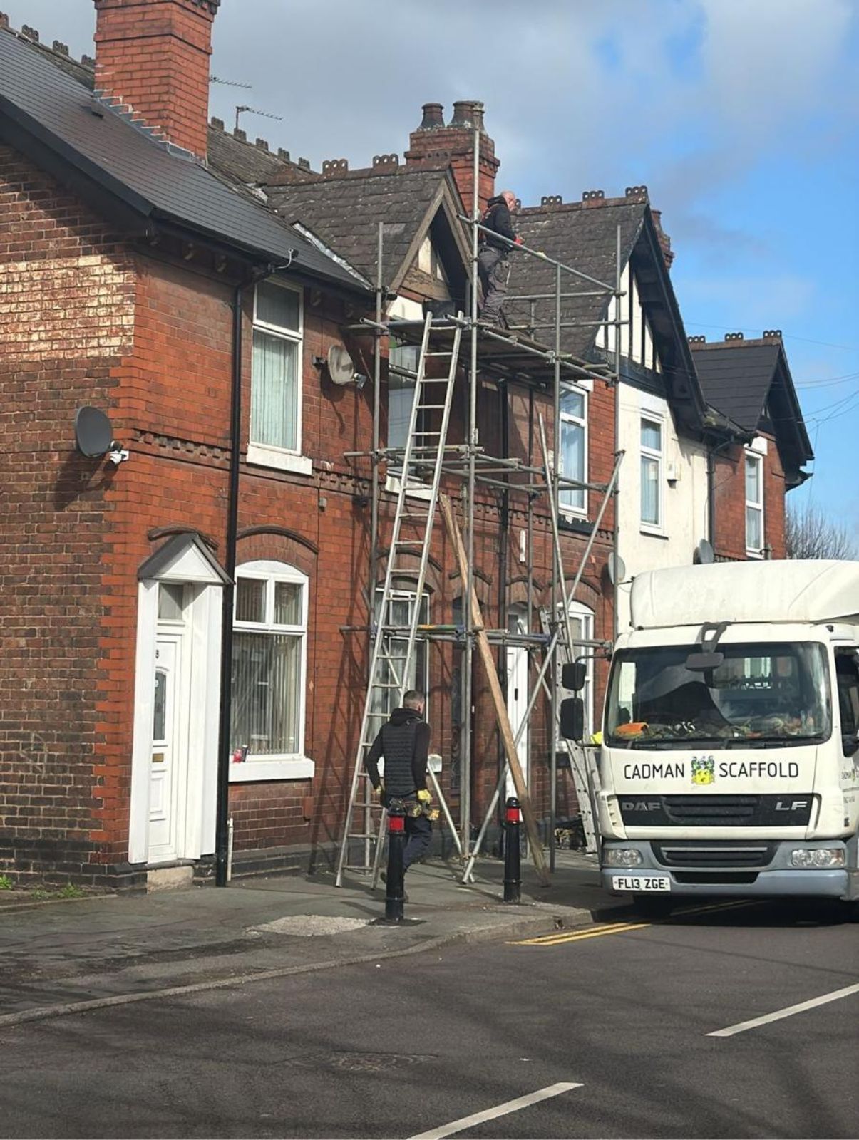 Workers on ladders repairing a brick house roof on a street, with a white van parked nearby.