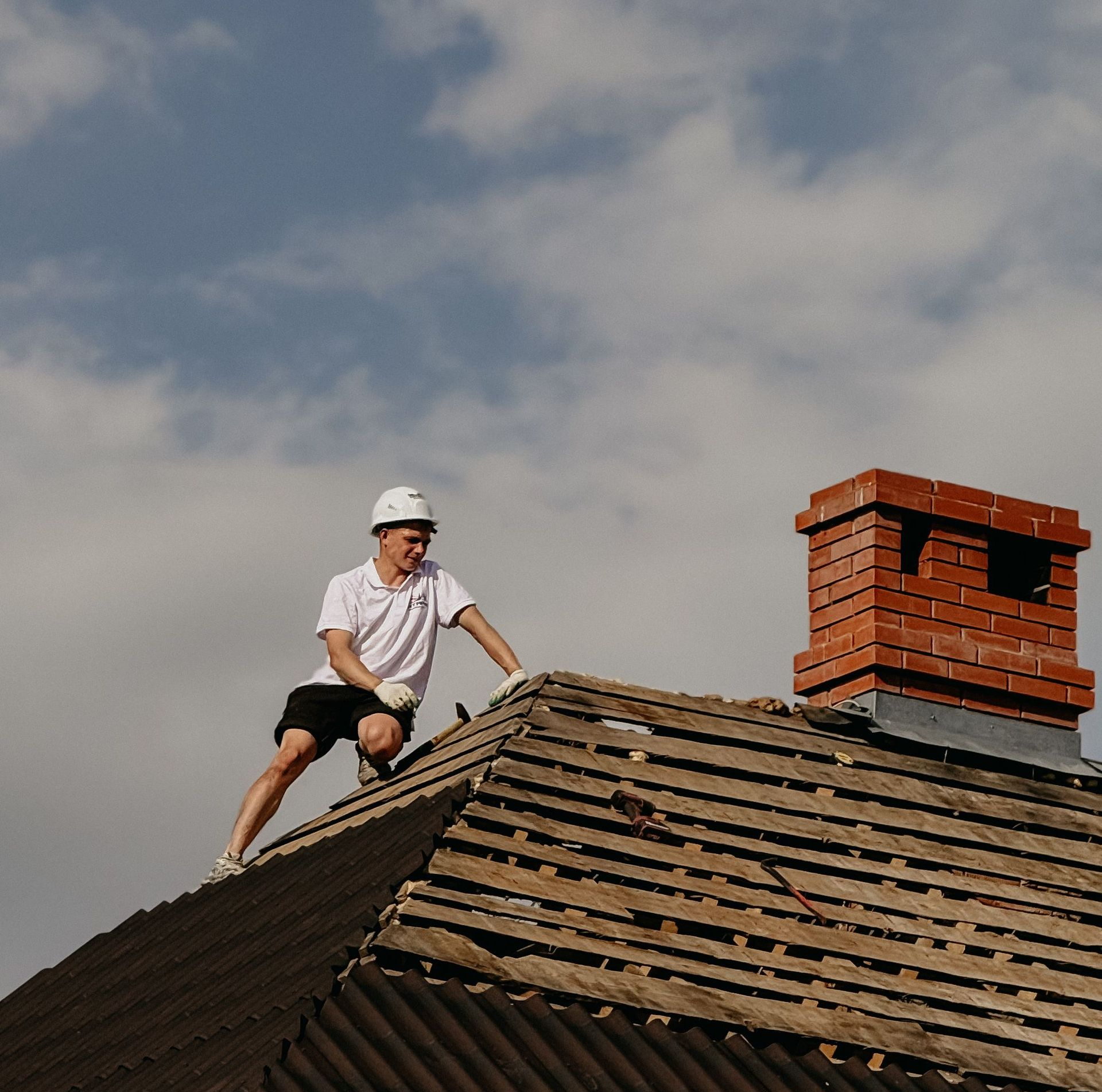 Worker in a white helmet repairing a shingled roof beside a brick chimney under a cloudy sky