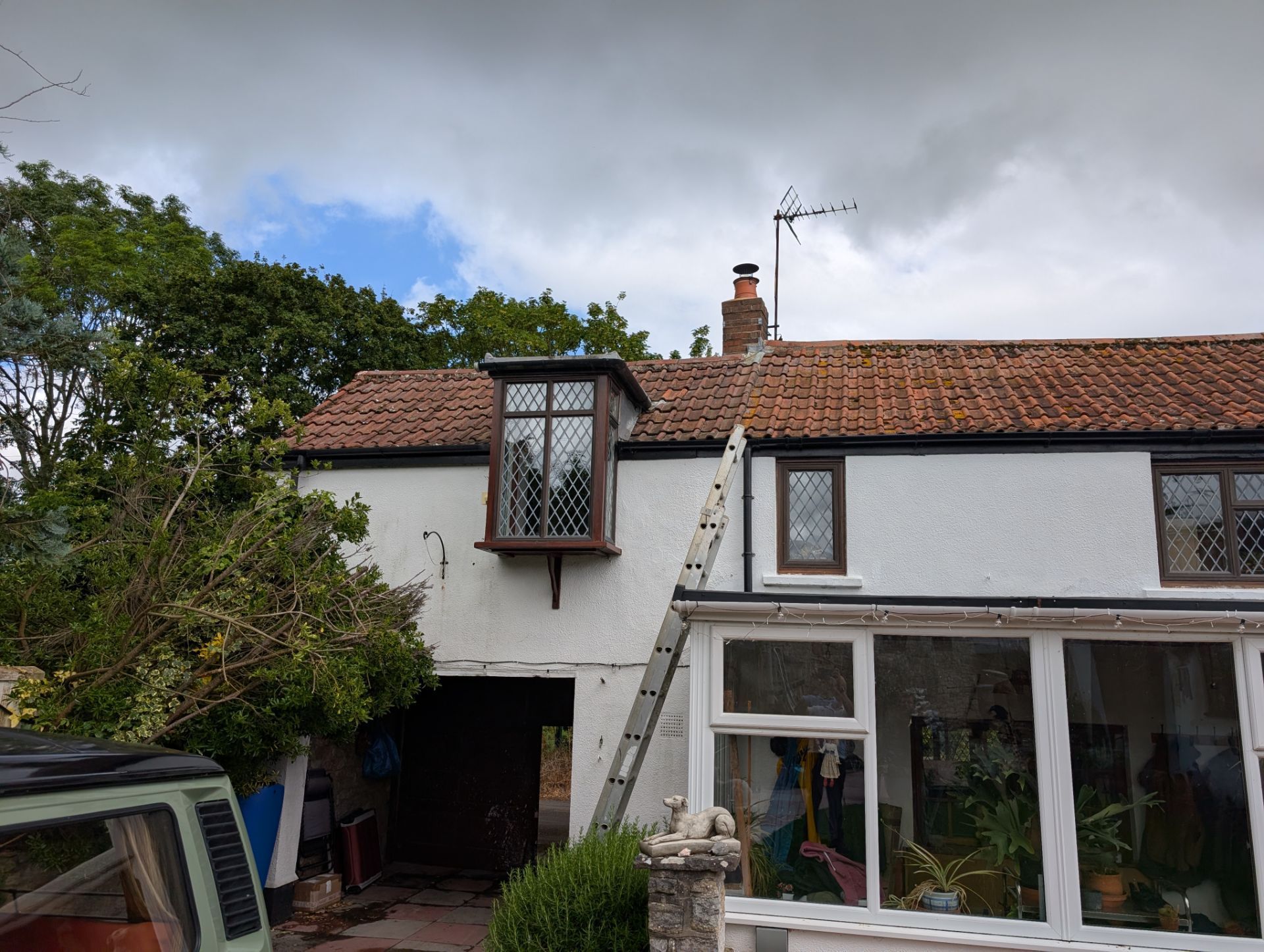 White house with red-tiled roof, ladder to an upper window, and a glass sunroom under a cloudy sky