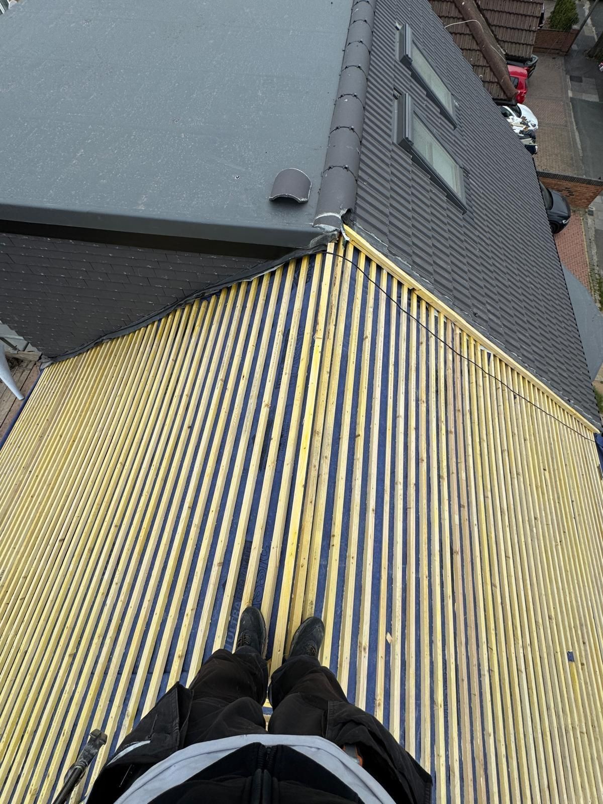 Looking down from a rooftop onto a yellow-striped sloped roof beside gray shingles and a brick wall