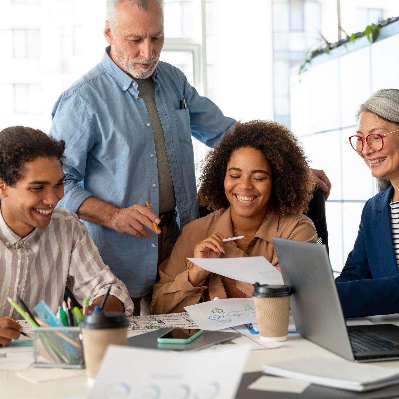 Office meeting: Diverse team smiles while reviewing documents, with a laptop and coffee cups.