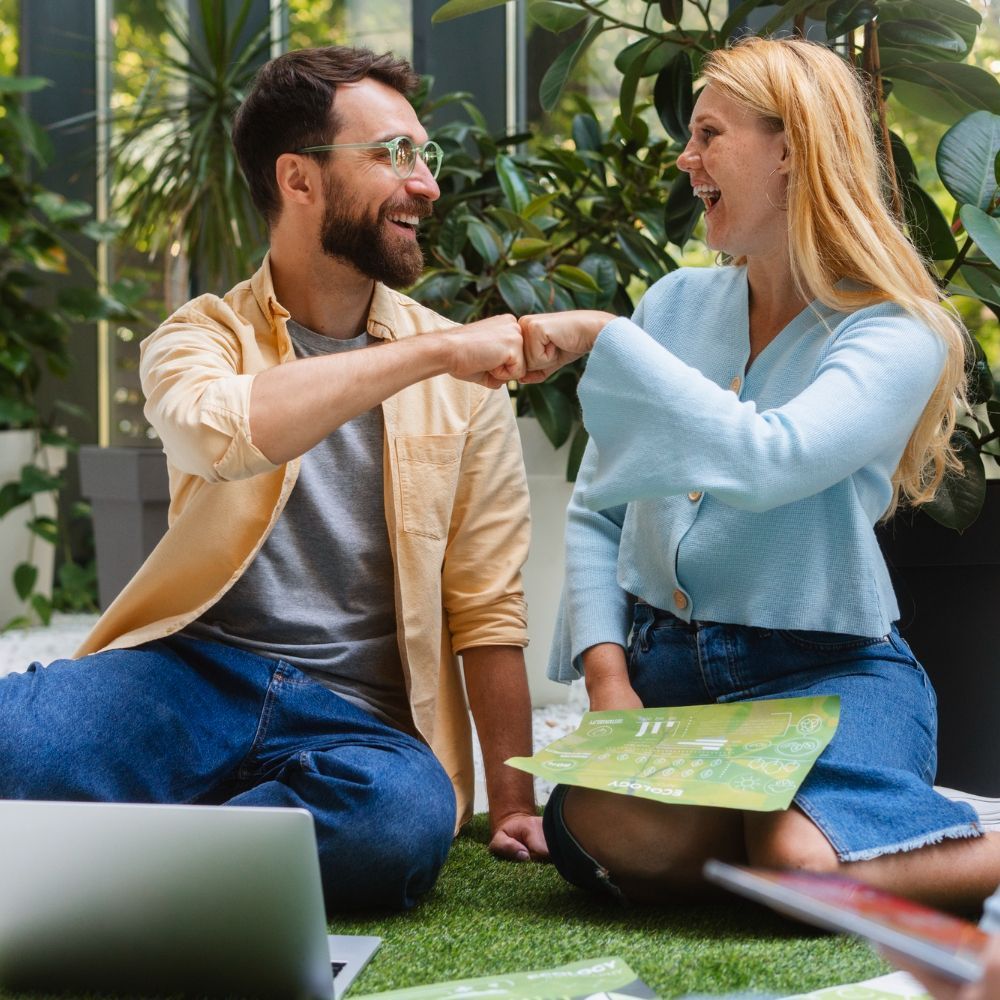 Man and woman fist bump, laughing outdoors near plants, a laptop, and papers.
