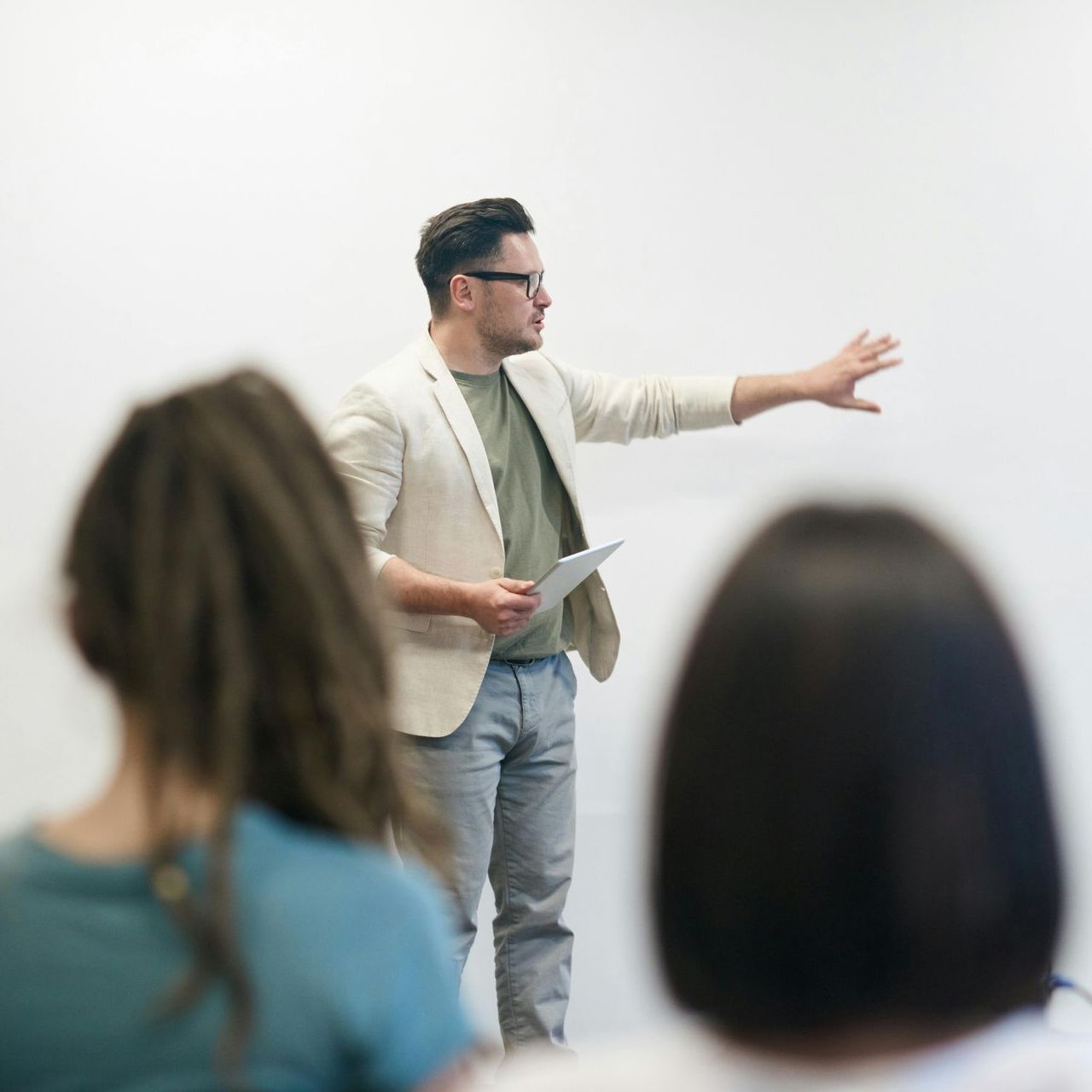 Man in a blazer lecturing, gesturing at white wall; two students in foreground.