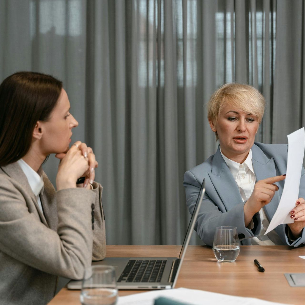 Two women in business attire at a table, one pointing at paper, the other thoughtful, with a laptop and water glasses.