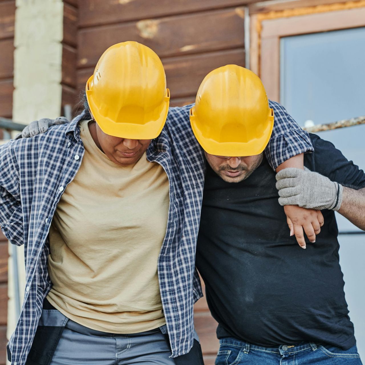 Construction worker helping injured colleague, both wearing yellow hard hats.