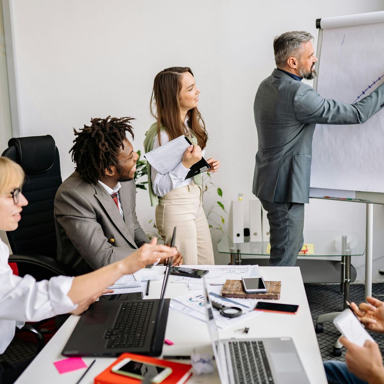 Business team in a meeting. A man writes on whiteboard, others watch, laptops on table.