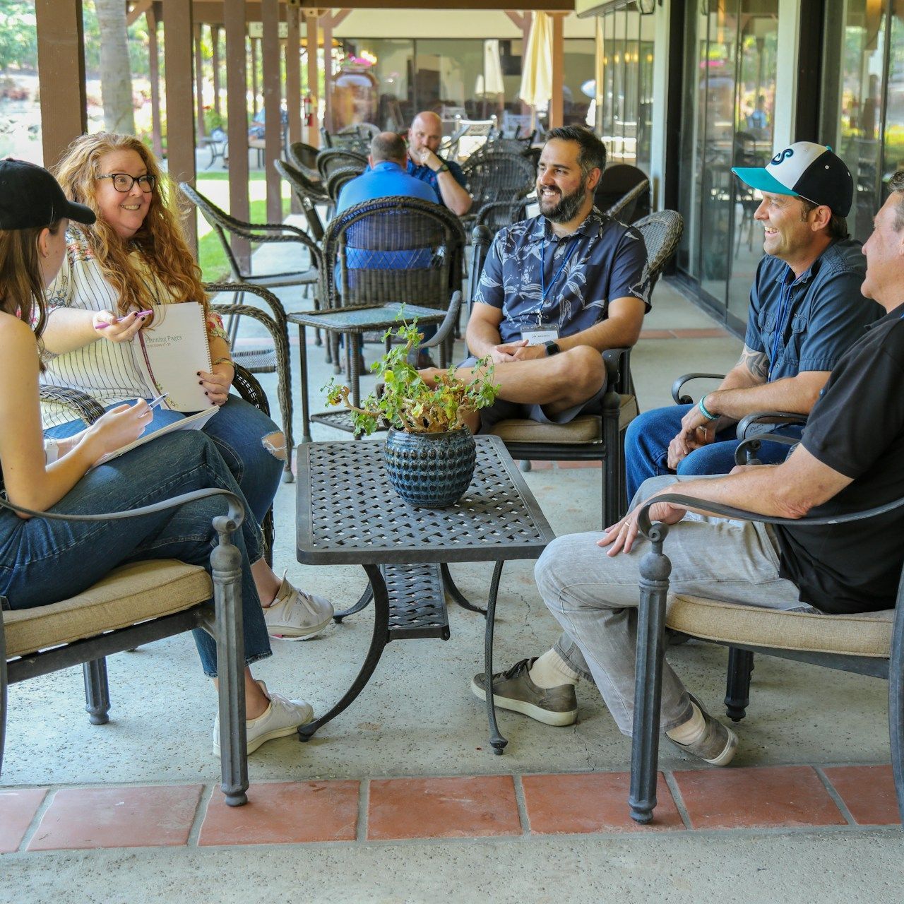 A group of six people sit outdoors, talking. Some are writing and smiling.