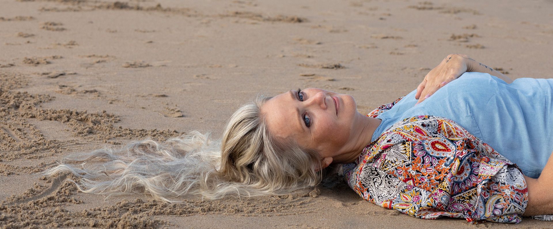 Vrouw ligt op het natte zand van het strand, kijkt opzij en draagt ​​een blauwe top met een kleurrijke kimono met patroon.