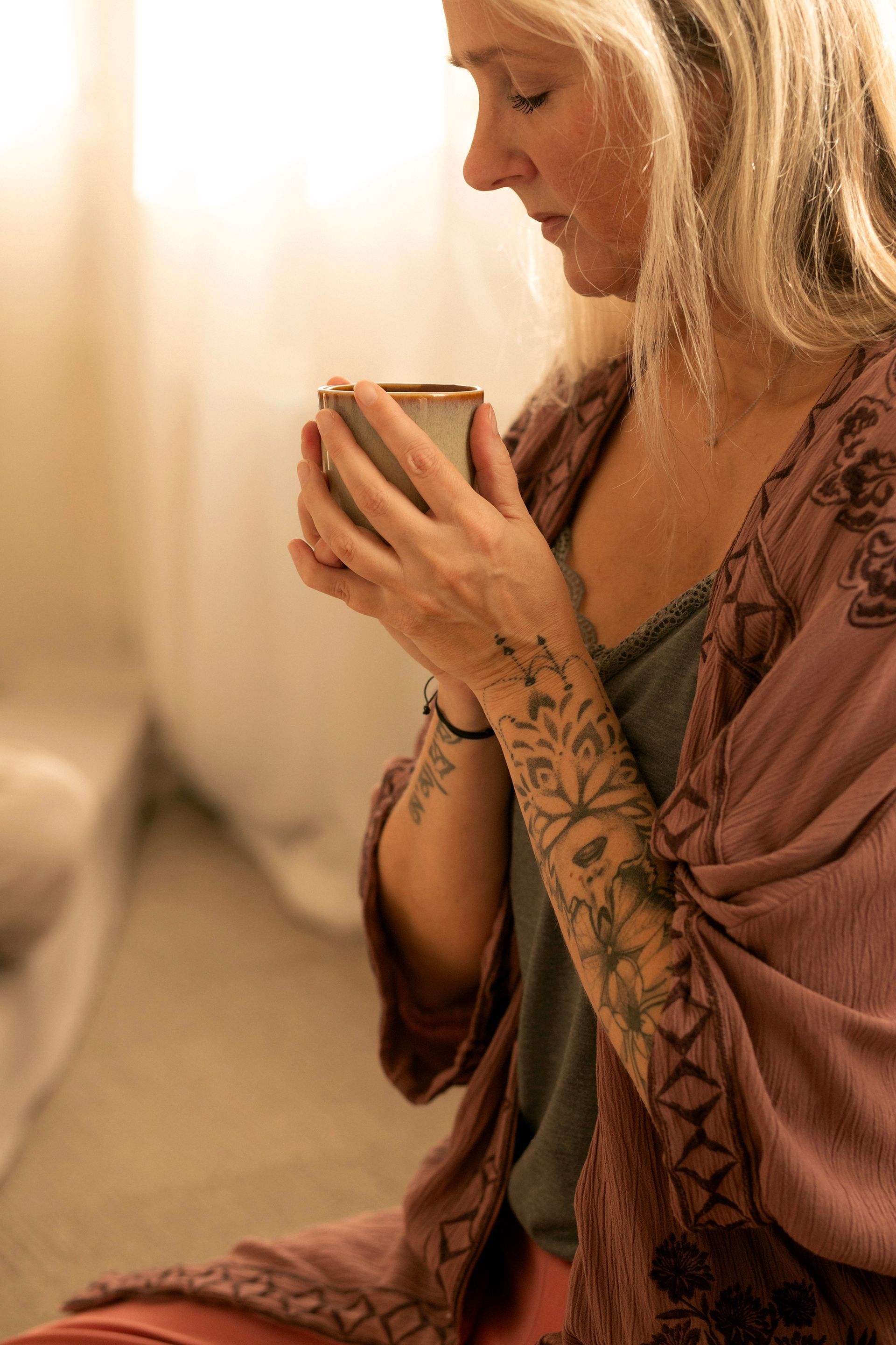 Een vrouw zit binnen, houdt een mok dicht bij haar gezicht en draagt ​​een mauve omslagdoek met henna-patronen op haar armen.