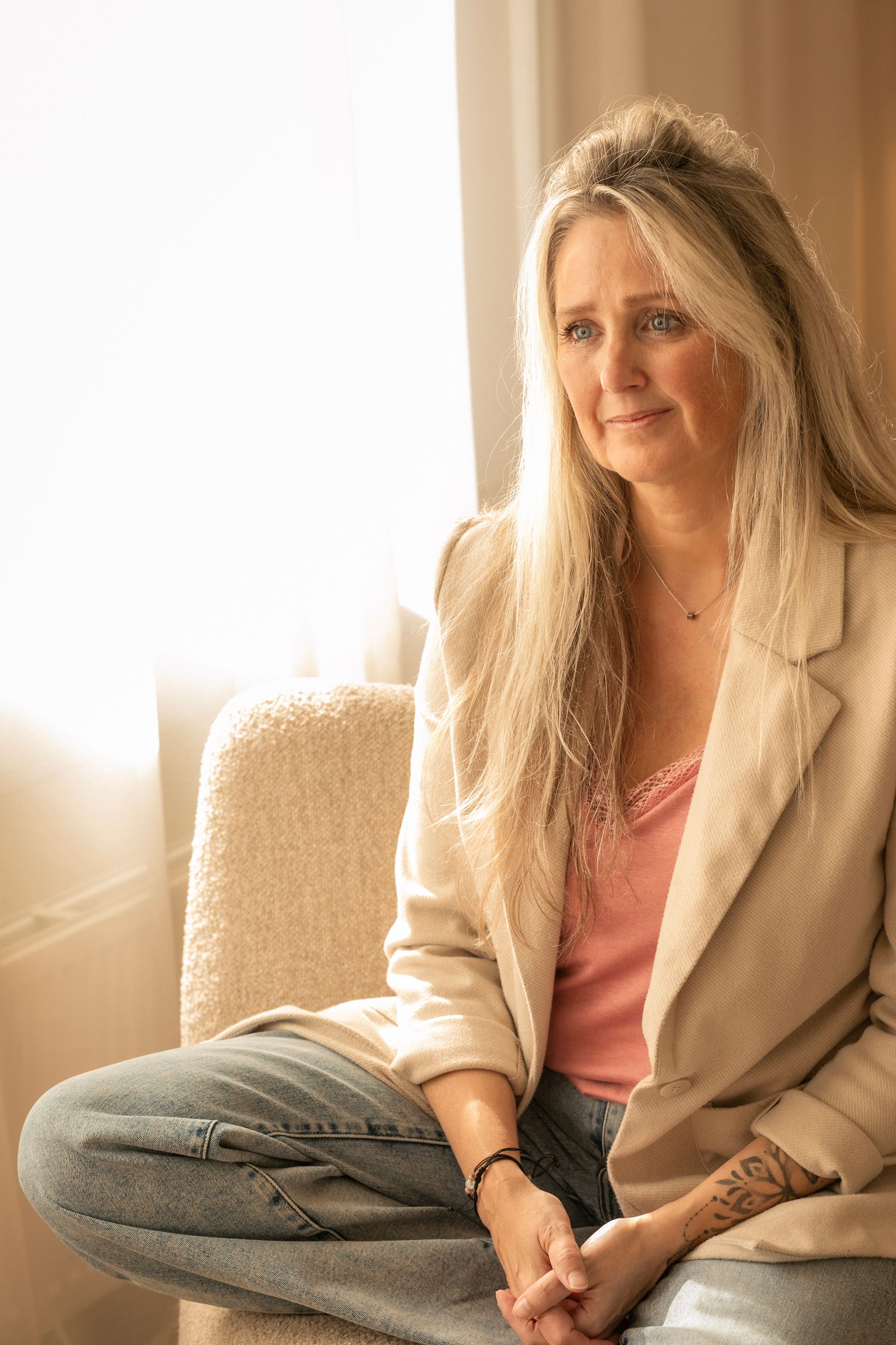 Een vrouw zit met gekruiste benen op een beige bank in een zonnige kamer, gekleed in een lichtgekleurde blazer en een roze top.