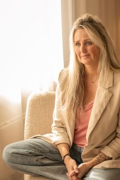 Een vrouw zit met gekruiste benen op een beige bank in een zonnige kamer, gekleed in een lichtgekleurde blazer en een roze top.