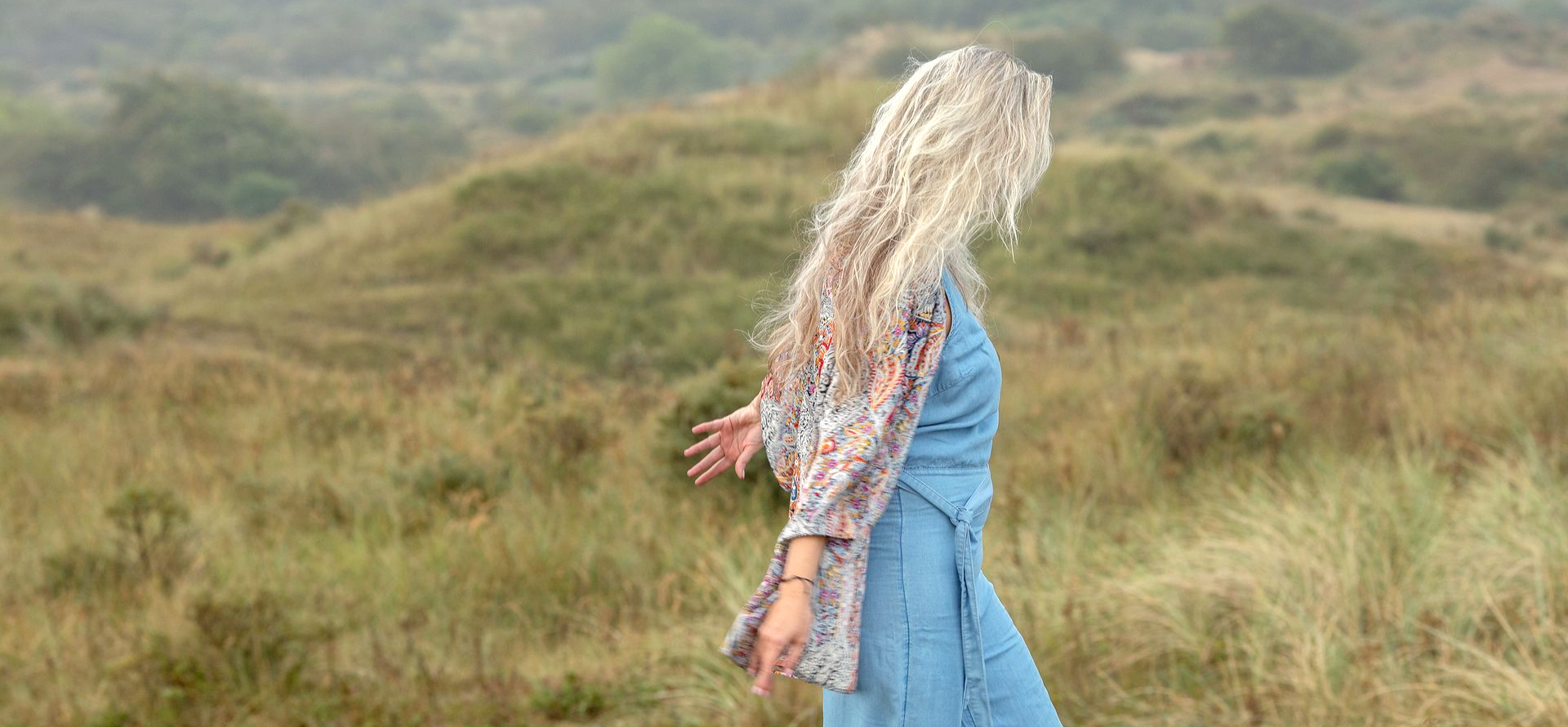 Een persoon in een blauwe jumpsuit met lang, licht haar loopt op de duinen onder een bewolkte hemel.