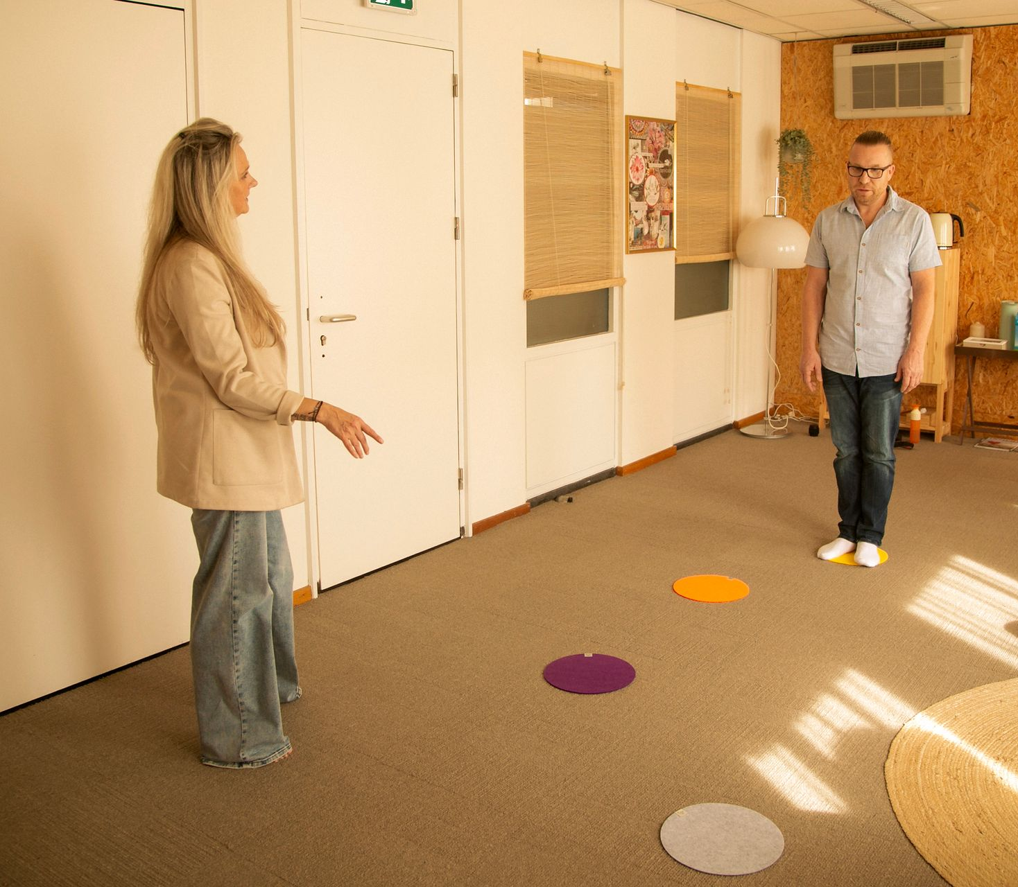 Twee personen zitten tegenover elkaar in een kamer met gekleurde vloermarkeringen voor een systemische oefening.