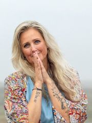 Vrouw met blond haar en tatoeages, glimlachend met de handen in een begroetingsgebaar tegen een effen achtergrond.