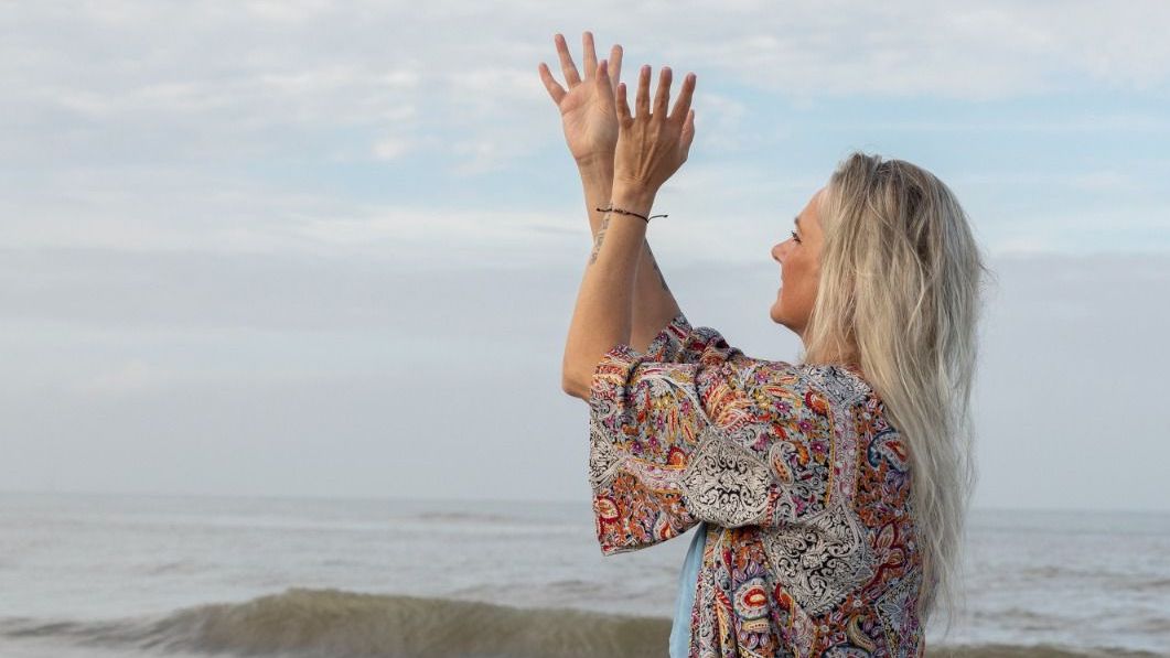 Persoon met opgeheven armen, kijkend naar de oceaan, op het strand, draagt een blouse met een patroon.