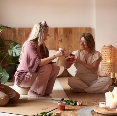 Twee mensen zitten op de grond en delen cacao in een knusse, bij kaarslicht verlichte kamer met planten en sfeervolle decoratie.