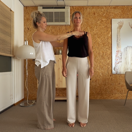 Twee vrouwen staan binnen; de ene meet de schouderbreedte van de andere in een kamer met houten lambrisering.