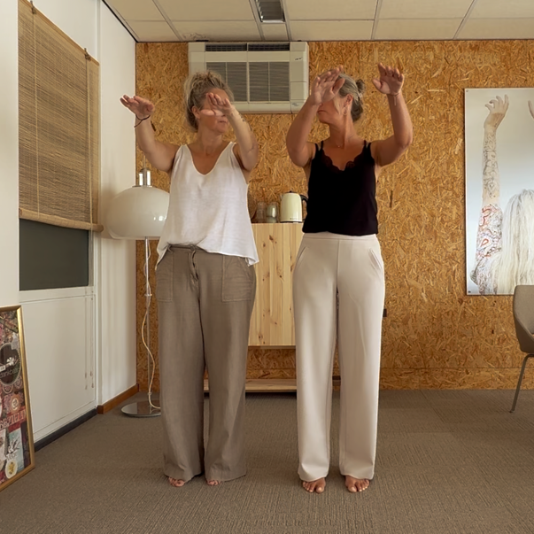 Twee mensen staan binnen met hun armen omhoog, in een kamer met houten wanden en een airconditioning.