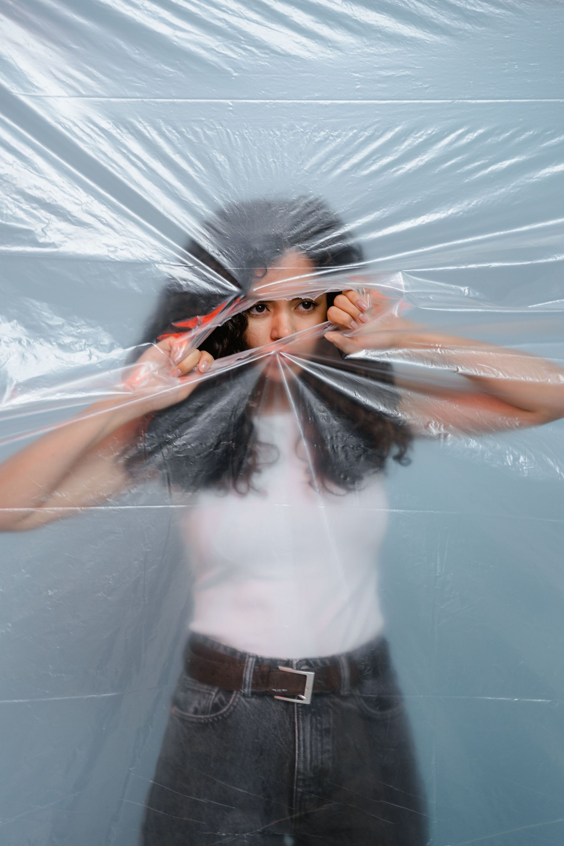 Person pressing hands against clear plastic film over face, standing against a light blue background.