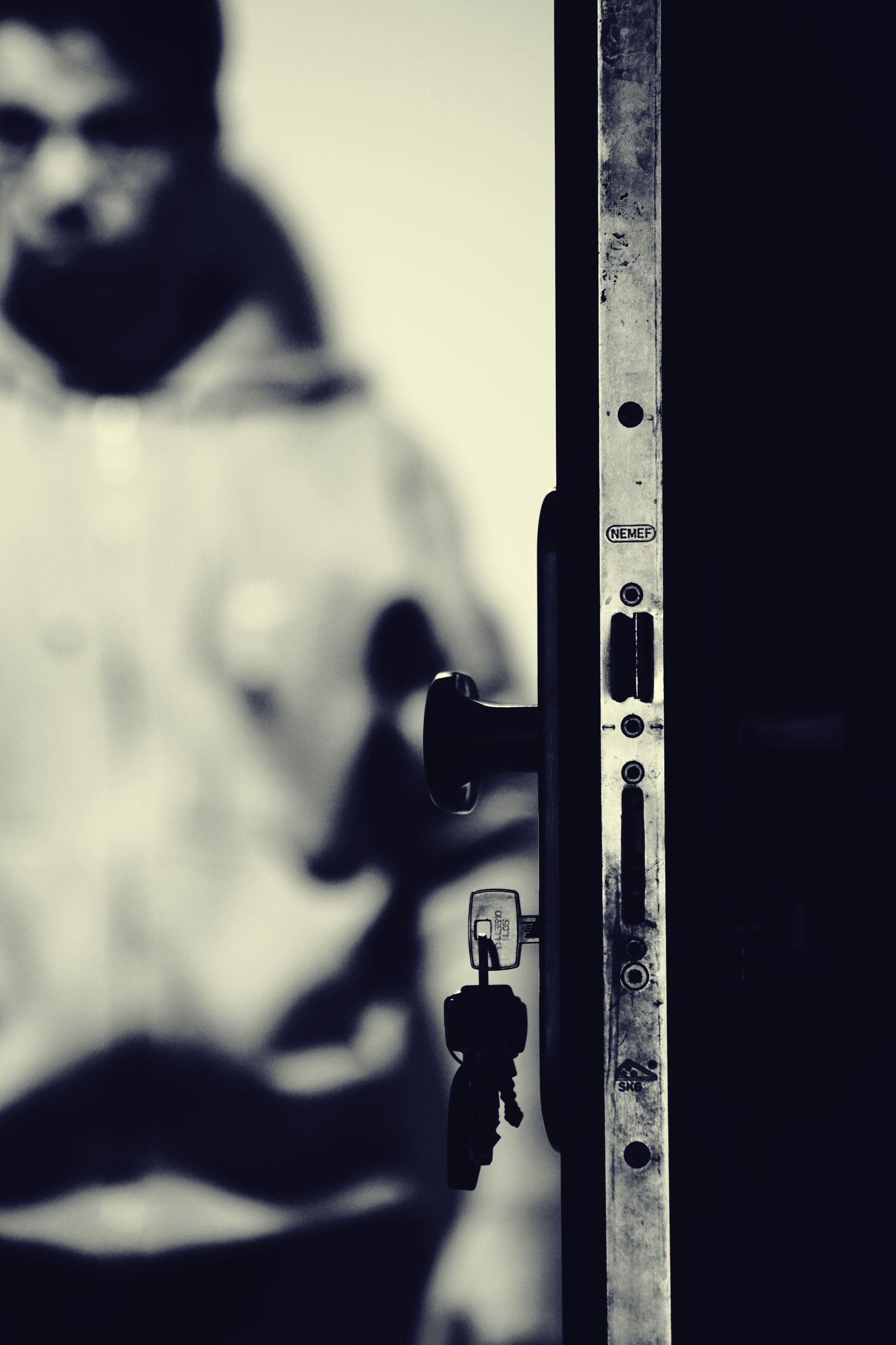 Blurred person in white beside a doorframe, with a small hanging lock in the foreground.