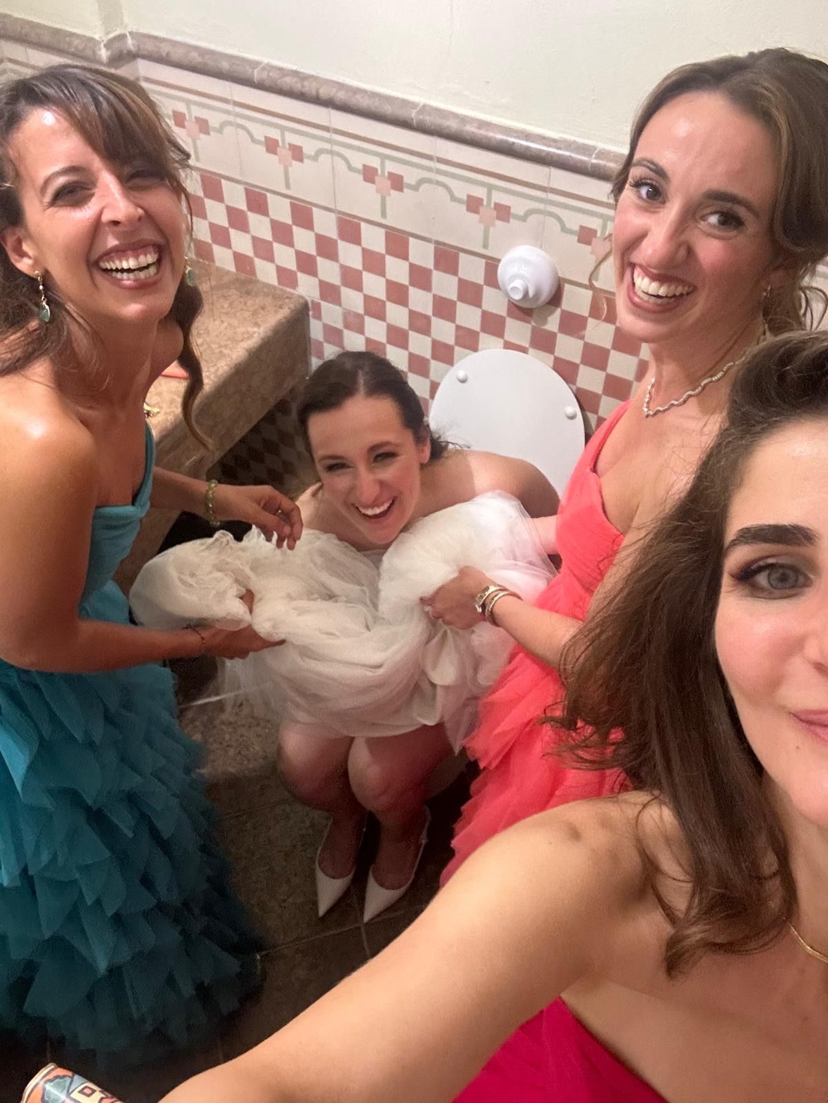 Four women in colorful dresses smiling in a bathroom, posing for a selfie around one in a white tulle dress.